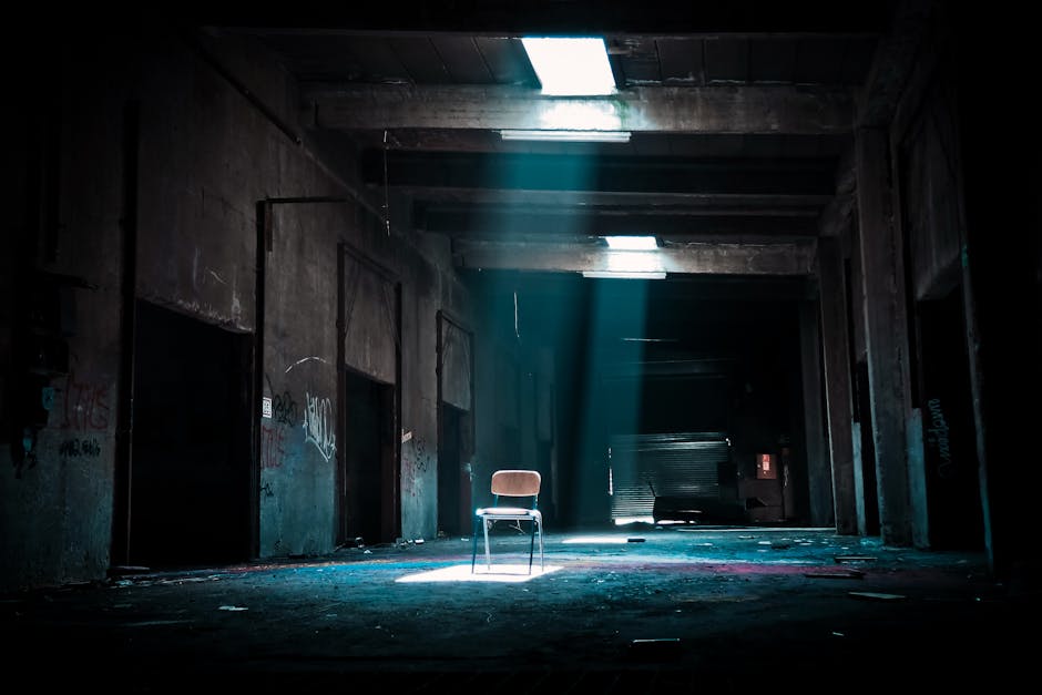 Eerie abandoned warehouse with a solitary chair illuminated by sunlight, evoking mystery.